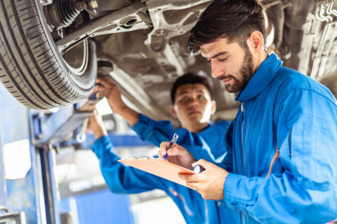 Two professional look technician inspecting car underbody and suspension system by using check list in modern car service shop. Automotive business or car repair concept.