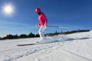 Young woman in pink winter jacket and helmet skiing snow spraying near, clear blue sky above