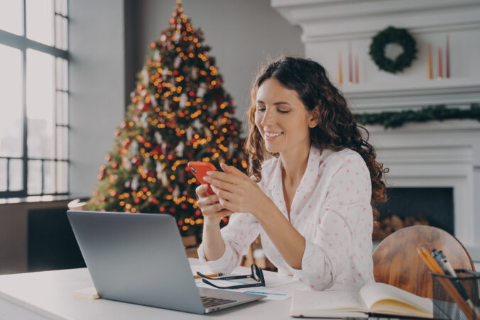 Joyful europian woman sitting at desk with laptop, reading xmas news or message on smartphone