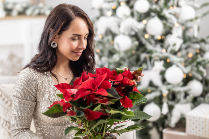 Woman smiles and smells a poinsette in a flower pot next to a Christmas tree.