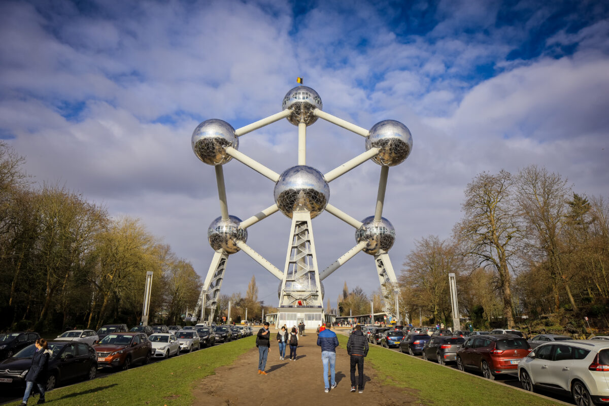 Visitors explore the Atomium in Brussels on a clear day among trees and parked cars in the foreground