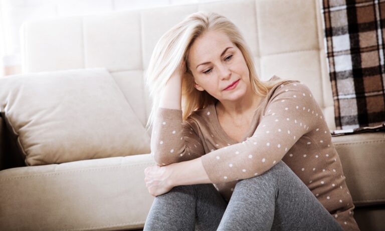Hormonálna terapia v menopauze. Žena v strednom veku premýšľajúca, sediaca na pohovke, symbolizuje obdobie menopauzy a rozhodovanie o hormonálnej terapii.