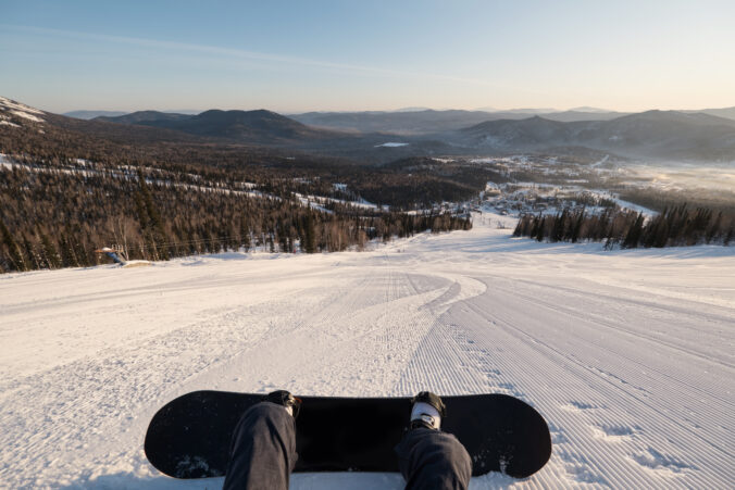 Female snowboarder lying on the snow on steep slope groomed and prepared for skiing, relaxing after riding, enjoying stunning view of winter mountains