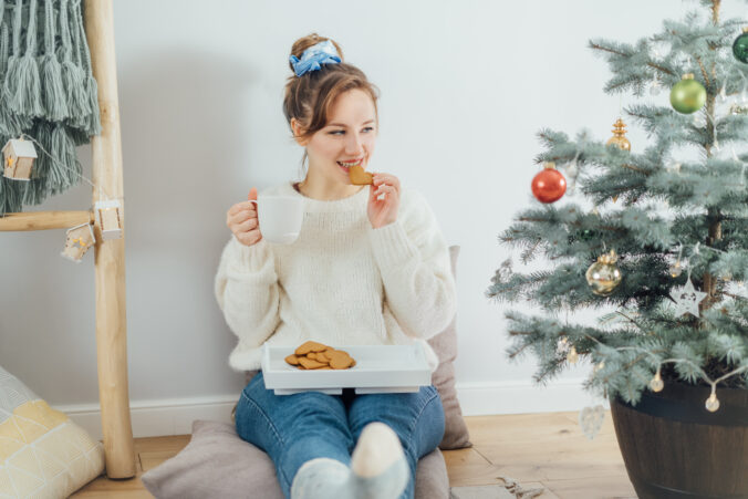 Smiling Young Woman in cozy sweater drinking hot cocoa with cookies, relaxing on the floor cushions near potted christmas tree in modern Scandi interior home. Eco friendly cozy winter holidays.