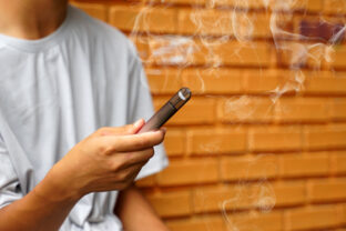 Asian male teenager holds a smoked e cigarette in his hand while sitting during recess in a hidden corner with his friends at school.