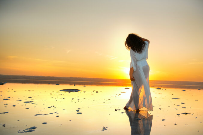 Beautiful girl with long dark curly hair in a long airy white dress stands in the water of a lake at sunset. mirror reflection of the sky in the surface of the lake. Fantastically beautiful view of a girl and nature