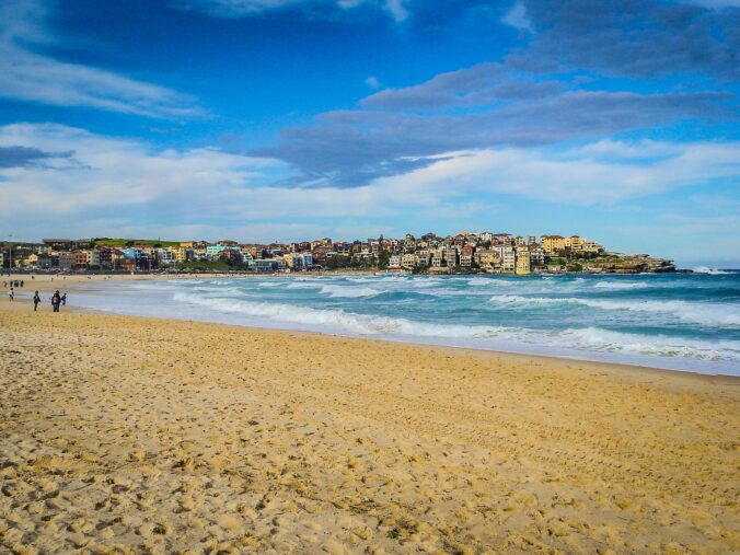 Pohľad na známu pláž Bondi v Sydney s jemným pieskom, vlnami oceánu a budovami na pozadí pod modrou oblohou.