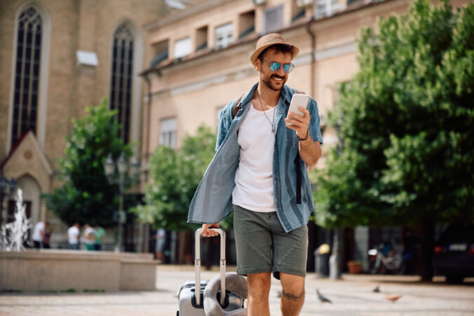 Happy man with travel bag text messaging on cell phone while walking in the city.