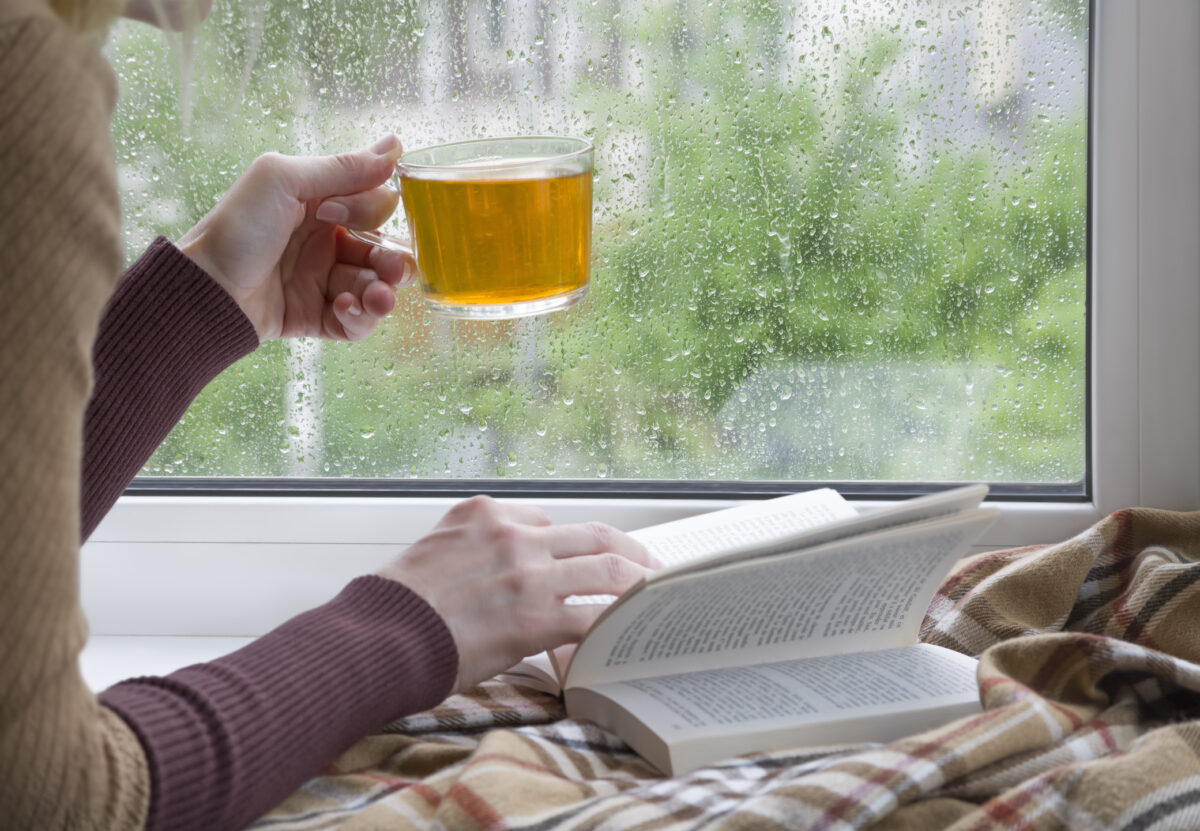 A woman's hand with a cup of tea. An open book and a soft blanket. The window glass is covered with raindrops. Springtime