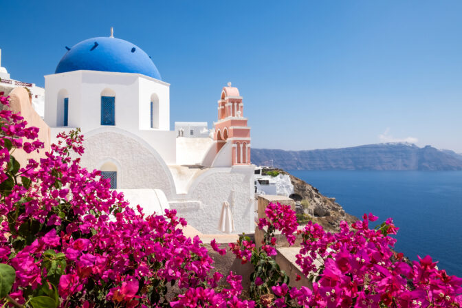 Santorini a sopka. Biela kupola a zvonica s výhľadom na modré more v tradičnej stredomorskej dedinke, obklopená ružovými kvetmi.