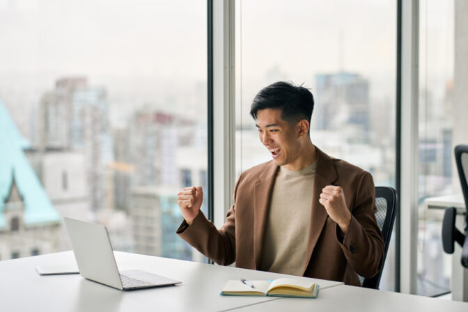 Happy Asian businessman celebrating success working in office looking at laptop.