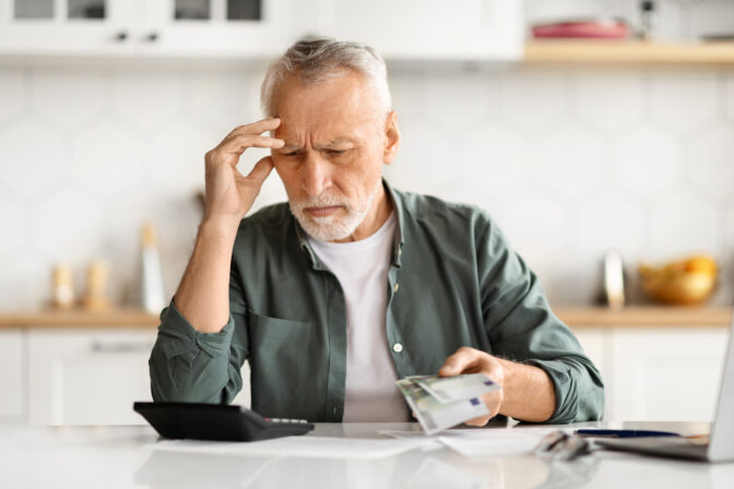Anxious senior man managing finances, holding cash and using calculator at home