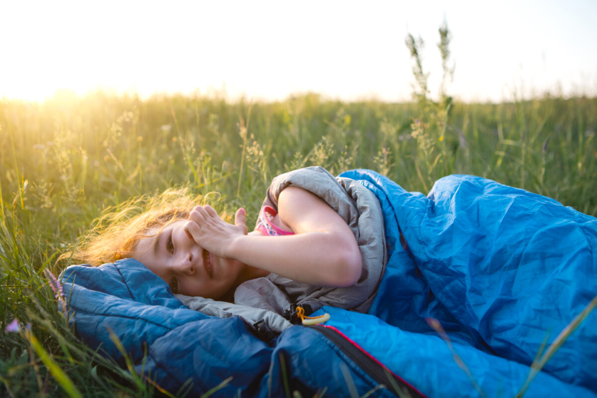 The girl is dissatisfied with scratching mosquito bites, child sleeps in a sleeping bag on the grass in a camping trip. Eco friendly outdoor recreation, summer time. Sleep disturbance, repellent.