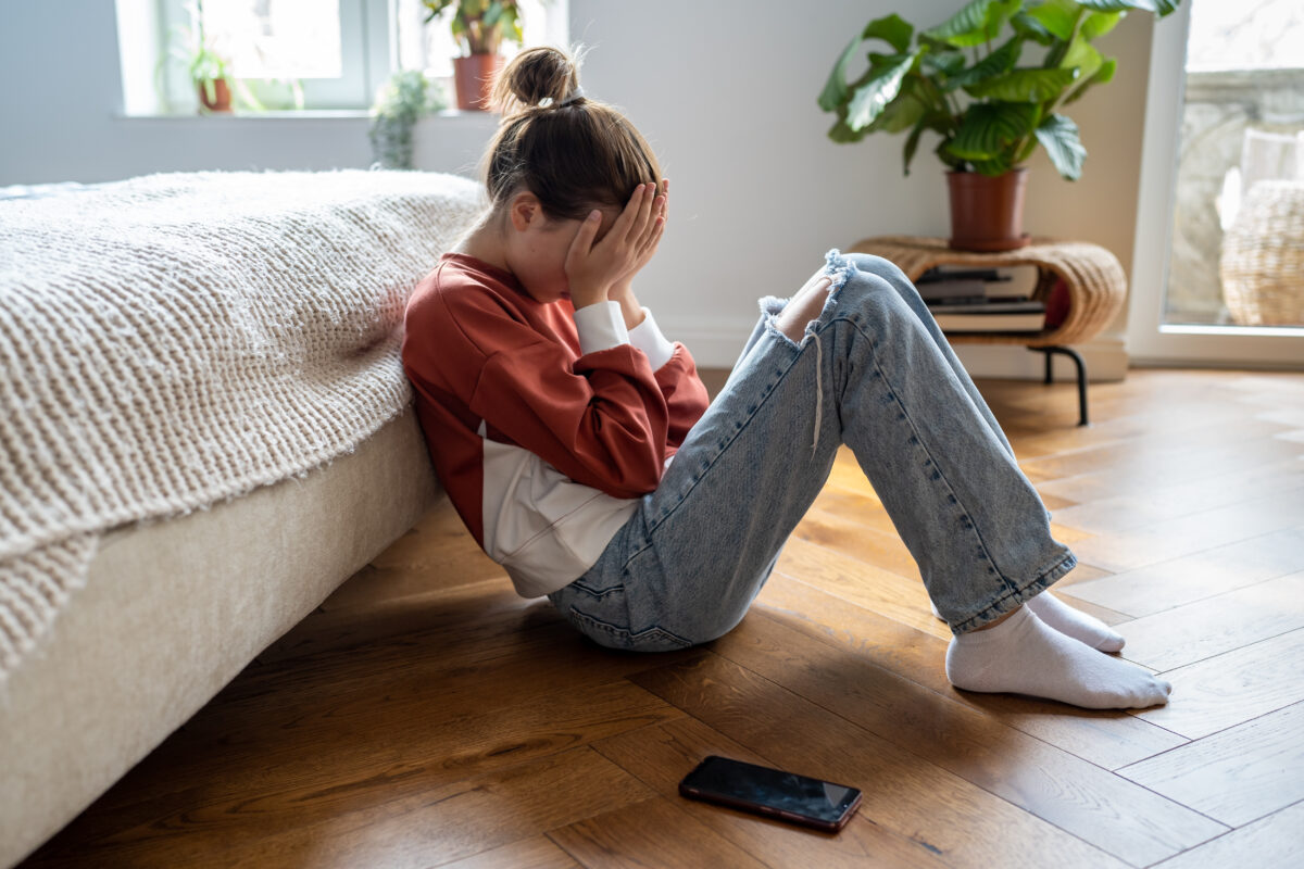 Unhappy teen girl covering face with hands crying while sitting on floor with mobile phone nearby