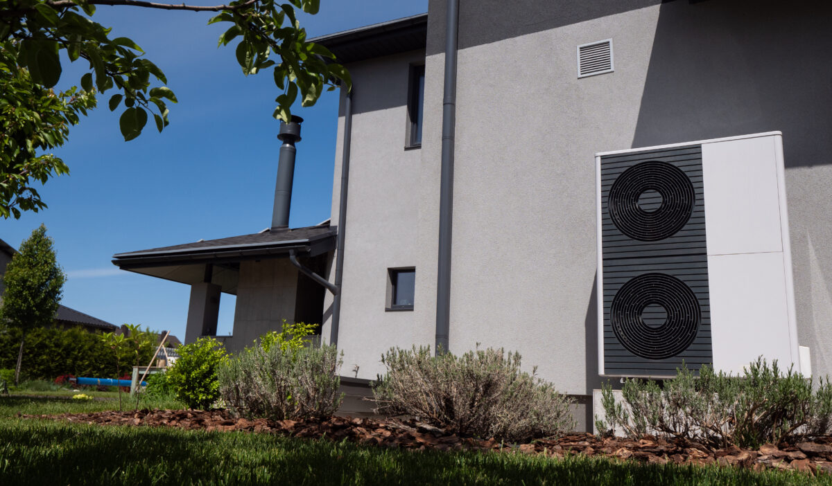 Positioned by the wall of a modern cottage, the outdoor unit of a heat pump is visible.