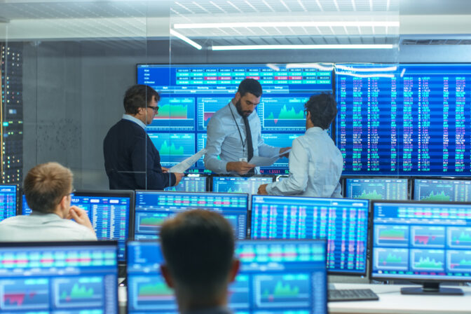 Three Experienced Stock Traders Talking Business, Consulting Documents and Argue About Data. They Work for a Big Stock Exchange Firm. Office is Full of Displays Showing Infographics and Numbers.