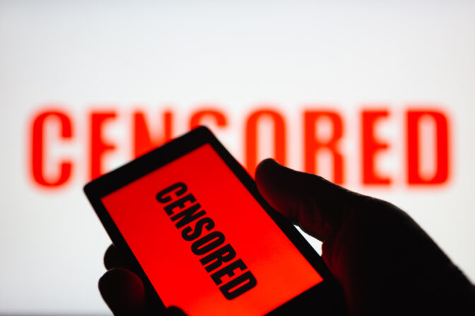 A smartphone in hand displaying the "censored" text. Same text in red blurred on white background. The concept of censorship on popular social networks. Restricted access to internet. Shallow DOF