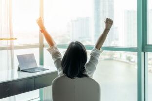 Business achievement concept with happy businesswoman relaxing in office or hotel room, resting and raising fists with ambition looking forward to city building urban scene through glass window