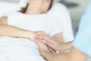 On bed lies a sick doctor sitting next to him and sympathetically holds hand