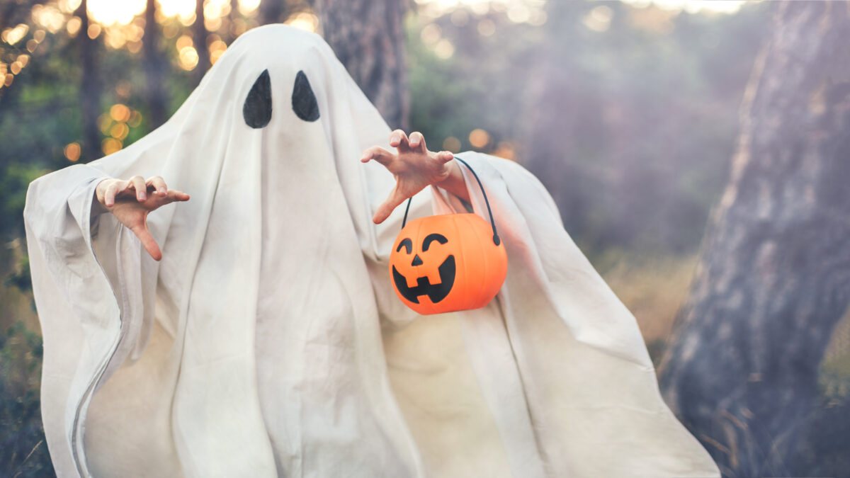 Girl wearing ghost costume holding pumpkin bucket with candies, standing in a forest.