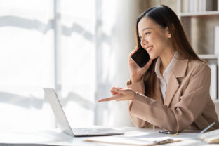 Attractive smiling asian businesswoman in light brown shirt talking on the phone with customer to name sales and checking finances on graph papers and laptop in office