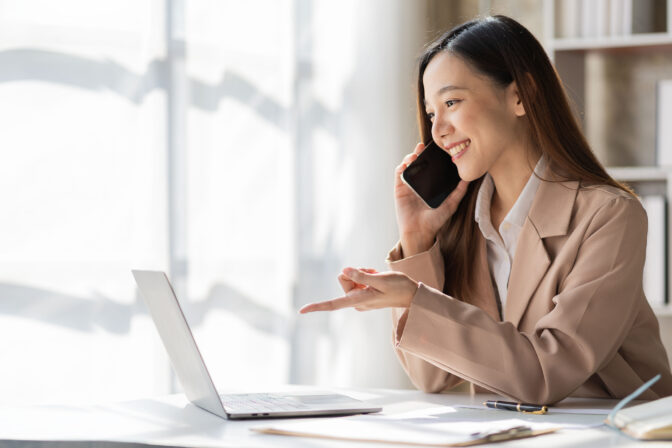 Attractive smiling asian businesswoman in light brown shirt talking on the phone with customer to name sales and checking finances on graph papers and laptop in office