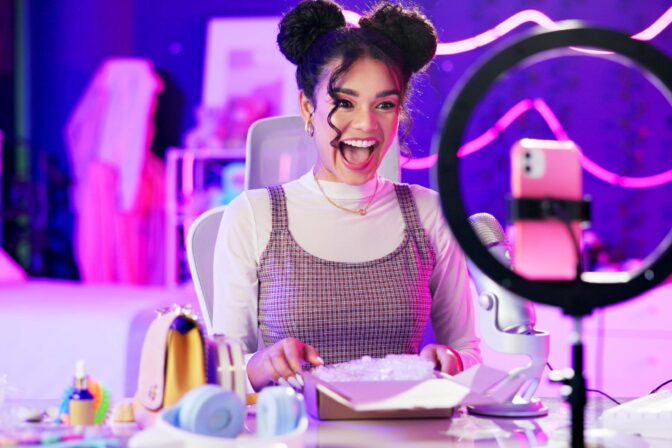 Unboxing, live stream and happy influencer woman with microphone, neon light and makeup in home studio. Content creator, beauty streamer or excited girl in online video for skincare product review