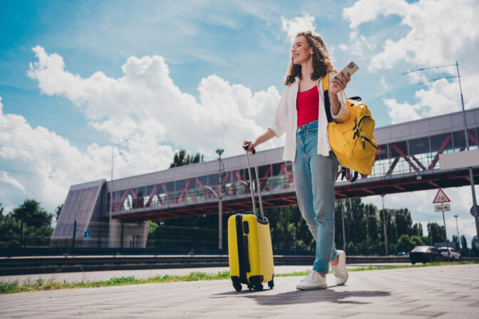 Full length photo of pretty dreamy lady tourist suitcase railway station arrival wear white shirt walking outside modern urban city street