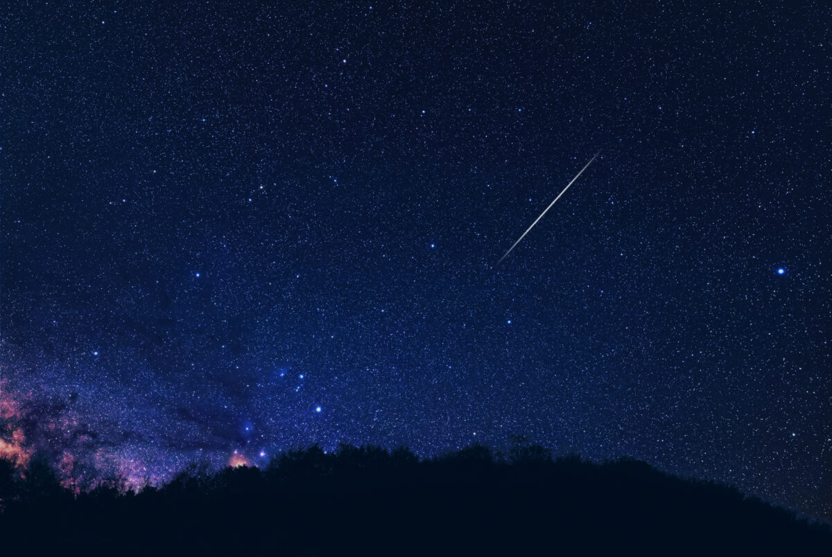 Milky Way stars with meteor shower trails and countryside tree silhouettes.