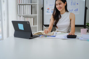 Young businesswoman analyzing financial documents, taking notes in a notebook, and utilizing a tablet for effective data management