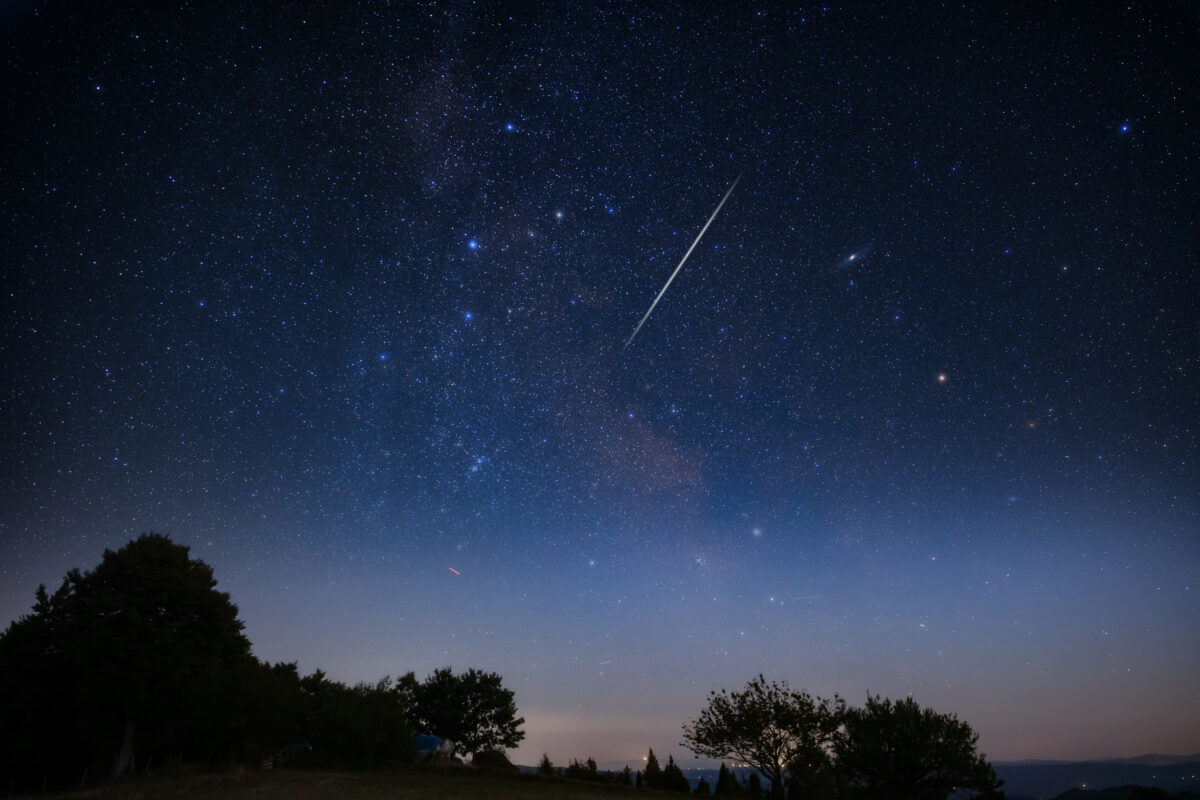 Milky Way stars with meteor shower trails and countryside tree silhouettes.