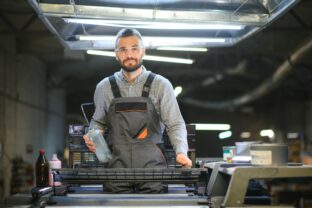 Printing house interior with modern offset print machine and operator in working uniform checking quality and controlling process of print