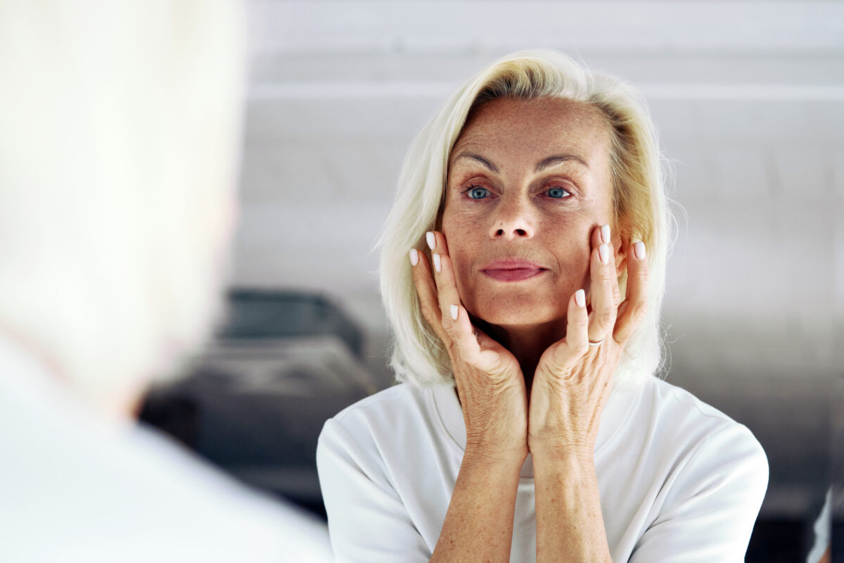 Older woman standing in front of mirror, gently touching her face with hands, close up