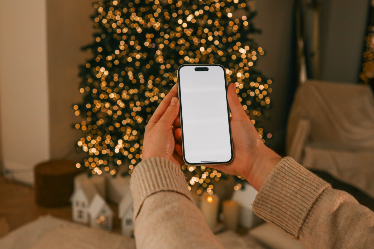Person holding a smartphone with a blank screen in a cozy holiday setting. A decorated Christmas tree sparkles warmly in the background, creating a festive and serene atmosphere
