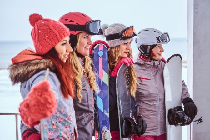 Women friends in sports winter clothes with snowboards and skis standing together at a snowy beach