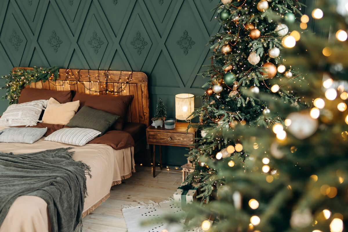 Bedroom decorated for Christmas. Bedroom interior with green walls, double bed and green Christmas tree decorated with emerald and bronze balls. View through blurred bokeh from garland lights. Nobody
