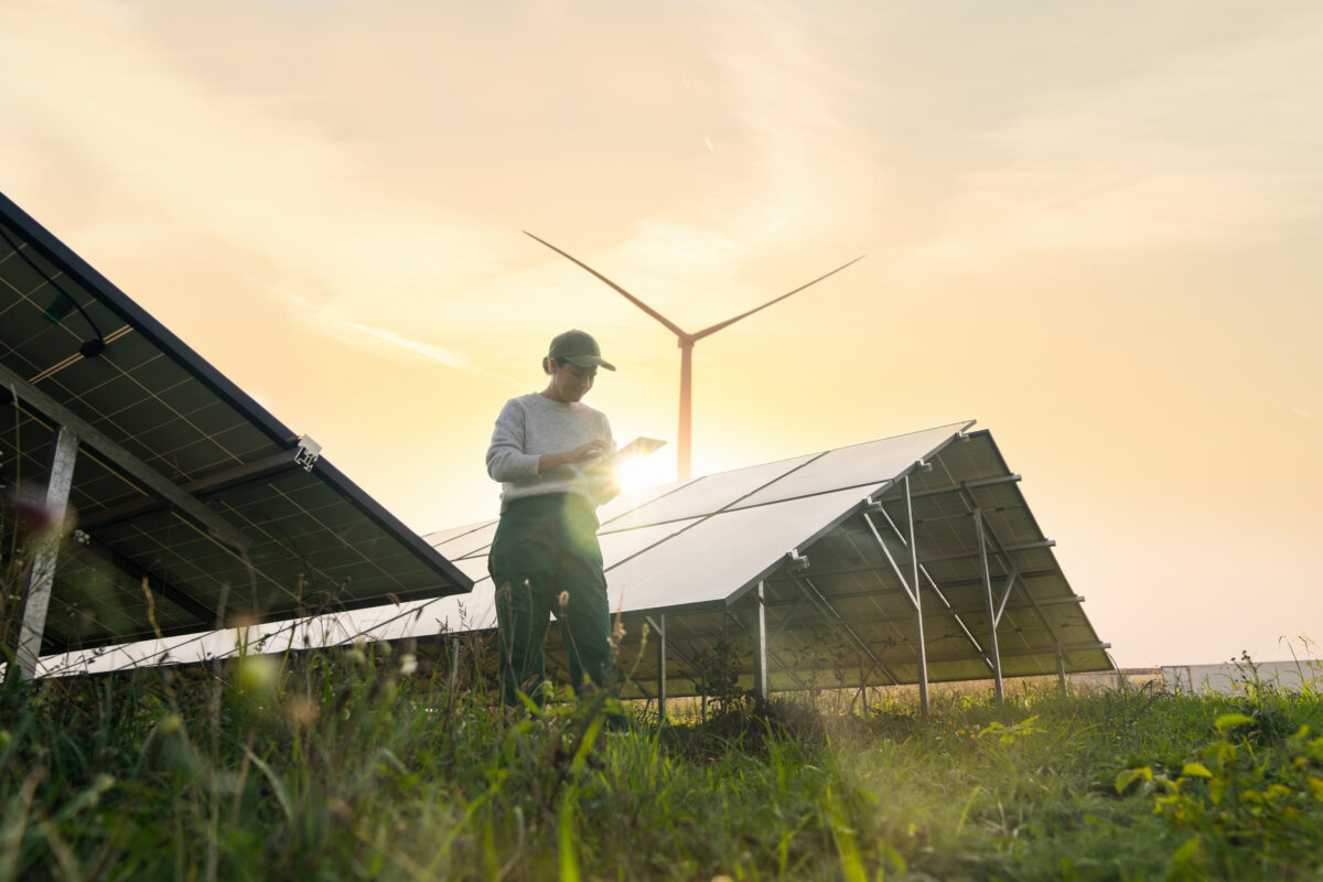 Female farmer wearing work uniform stands next to solar panels. Modern agricultural farm using solar panels. Agricultural silos in the background.