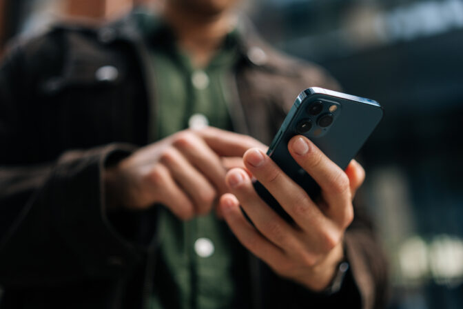 Close up hands of unrecognizable man holding and using smartphone standing on city street, browsing internet, checking social media, using mobile application.