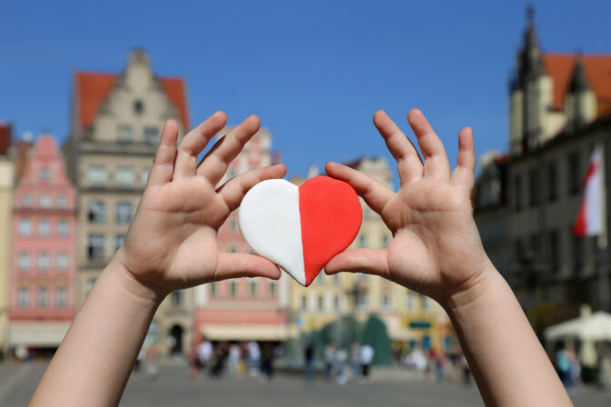 A red white heart in the colors of the national flag of Poland in the hands of a child against the backdrop of the old town square. Independence Day of Poland. Freedom and Democracy