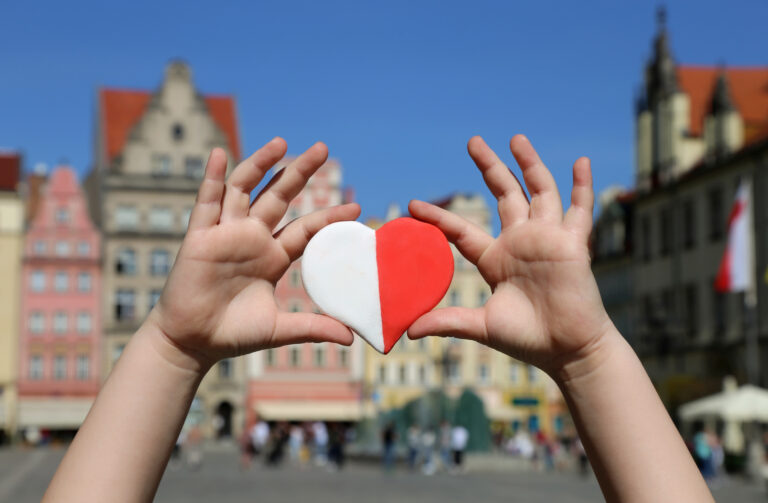 A red white heart in the colors of the national flag of Poland in the hands of a child against the backdrop of the old town square. Independence Day of Poland. Freedom and Democracy