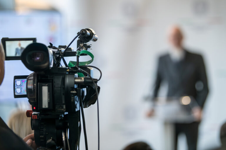 TV camera filming live press event with officials at podium. Highlights media coordination, lighting precision, and structured public communication.