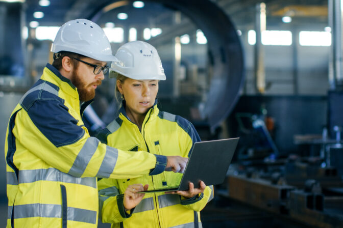 Male and Female Industrial Engineers in Hard Hats Discuss New Project while Using Laptop. They Make Showing Gestures.They Work in a Heavy Industry Manufacturing Factory.