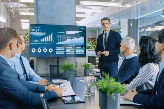 Young Stock Trader Shows to the Executive Managers Cryptocurrency and Trade Market Correlation Pointing at the Wall TV.