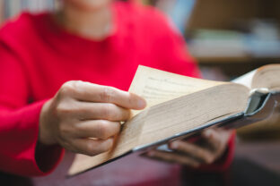 Person in vibrant red sweater holds an open book, delicately turning page, embodying joy of reading and learning in warm, inviting environment
