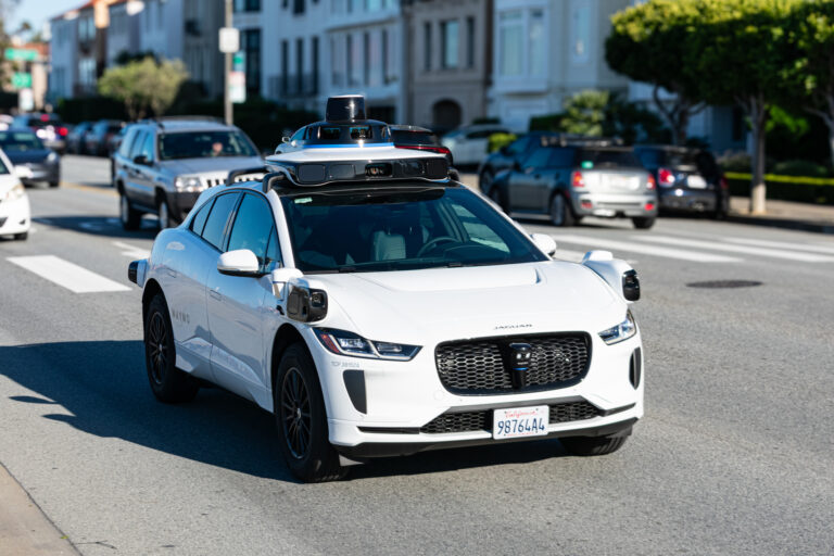 Waymo One Jaguar driverless autonomous car equipped with lidar sensors and cameras drives on a multi lane city road. Front side view. Selective focus