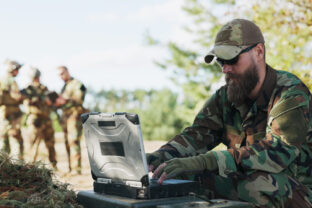 Soldiers stand ready in the field, dressed in full gear with weapons in hand, awaiting orders while their commander transmits tactical data to the base from a computer.