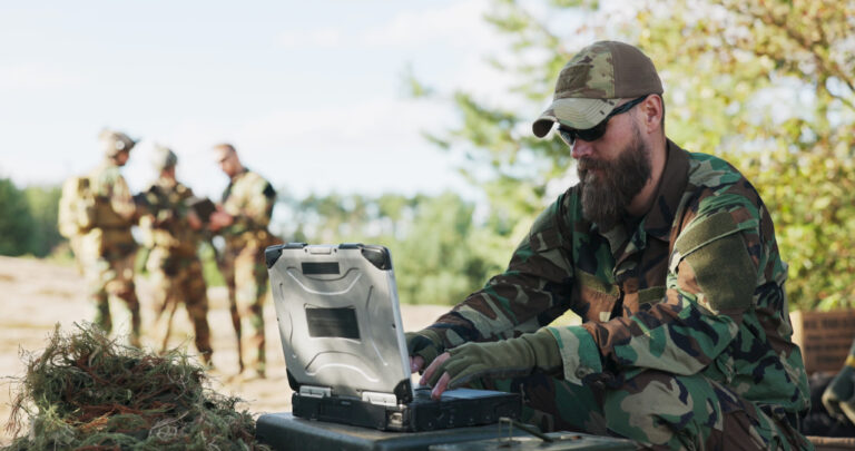 Soldiers stand ready in the field, dressed in full gear with weapons in hand, awaiting orders while their commander transmits tactical data to the base from a computer.