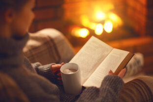 Book and cup of coffee in hands of girl on  winter evening near fireplace