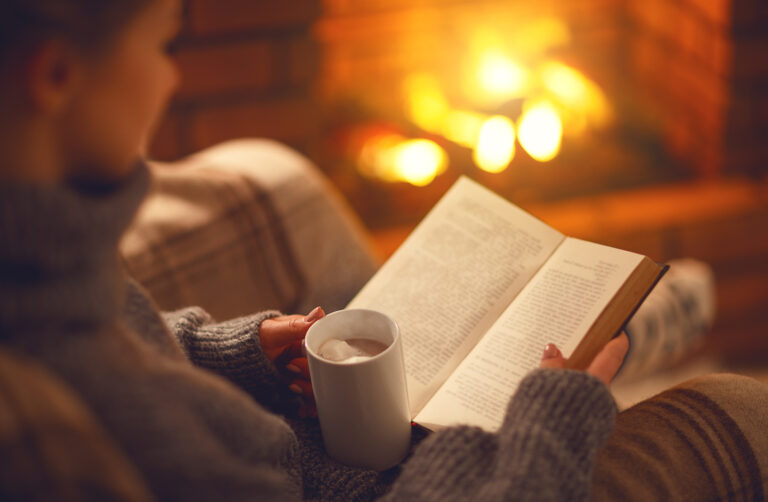 Book and cup of coffee in hands of girl on  winter evening near fireplace
