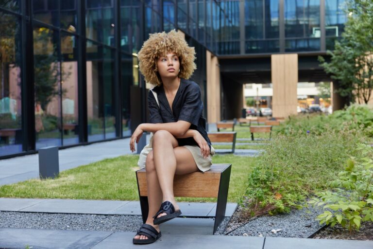 Outdoor shot of pretty millennial woman with curly hair dressed in casual wear poses on wooden bench against urban background looks thoughtfully somewhere rests after strolling at business centre